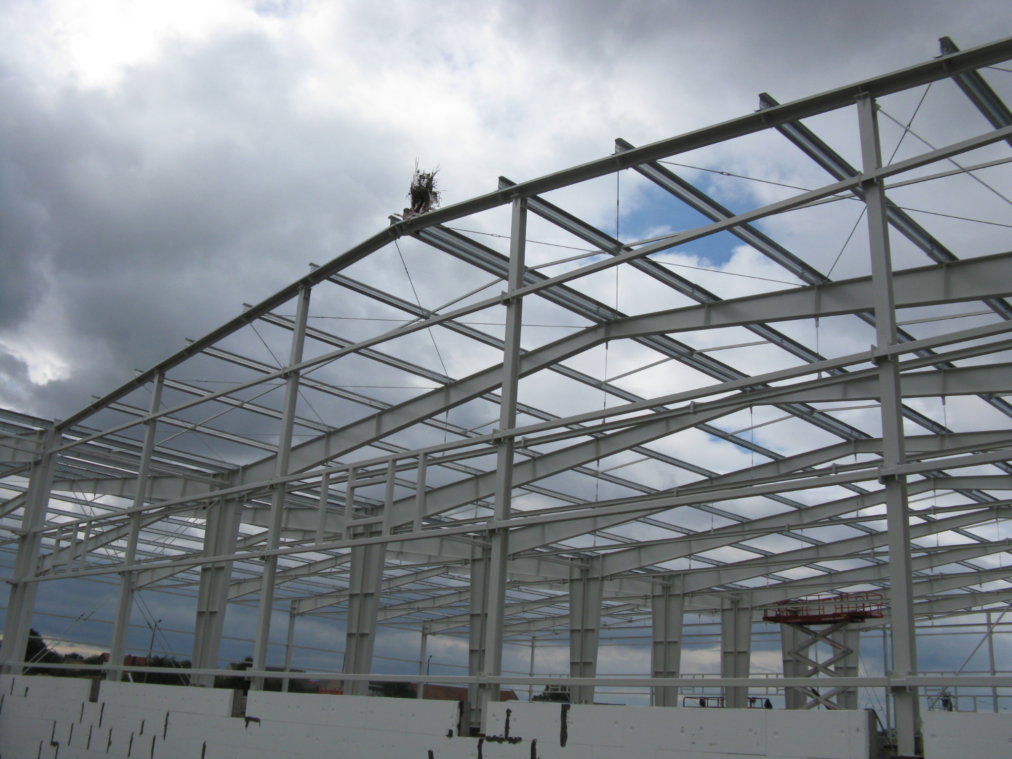 Wnętrze budowanej hali stalowej z widoczną konstrukcją szkieletową i podnośnikiem koszowym, na tle pochmurnego nieba z prześwitami błękitu.