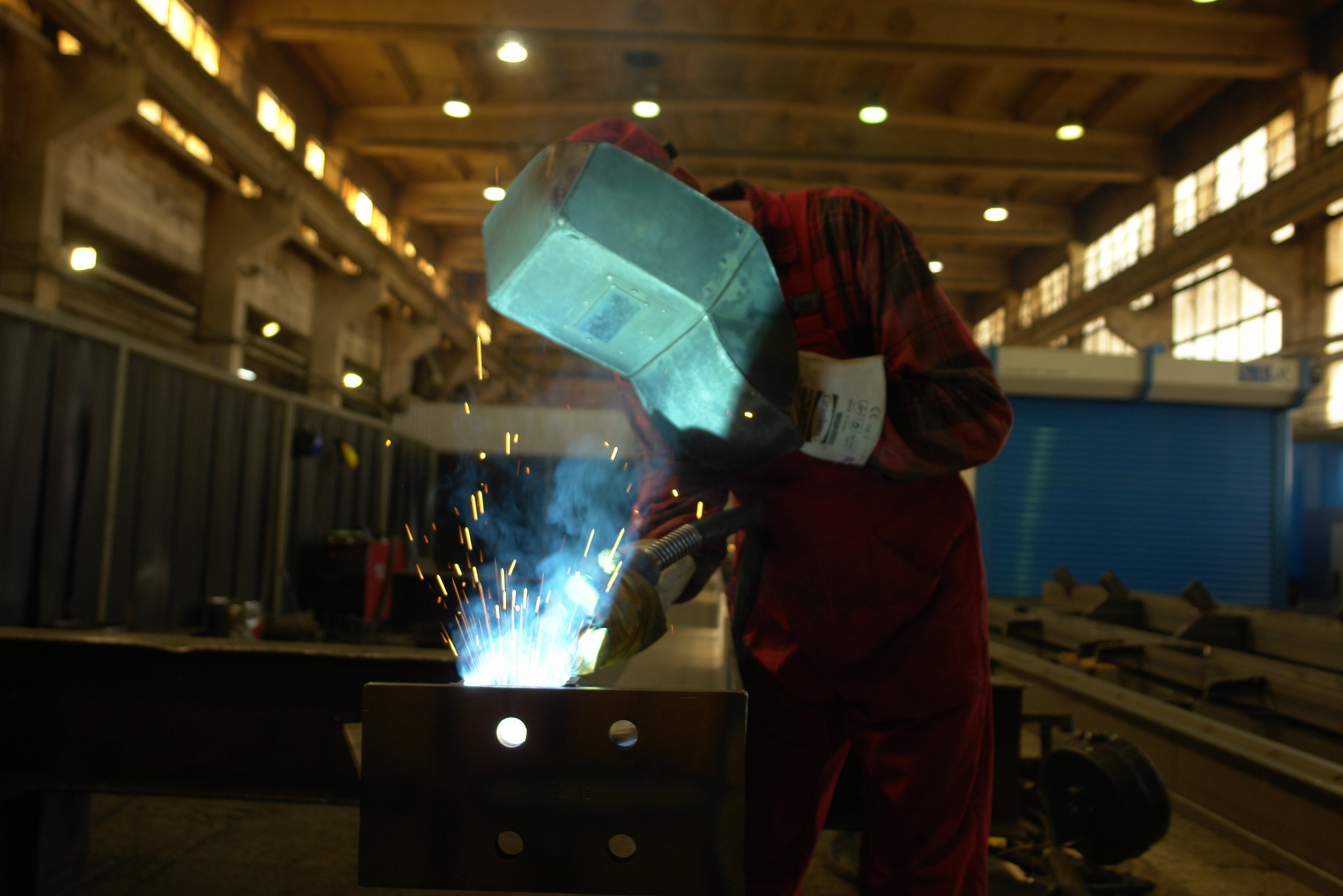 Spawacz w czerwonym kombinezonie i masce spawalniczej łączy metalowe elementy konstrukcji, iskry i dym podczas spawania, widoczne oświetlenie hali.