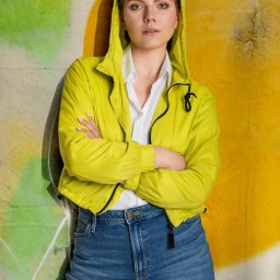 Studio Detal - Portret kobiety w limonkowej kurtce z kapturem, opierającej się o ścianę z graffiti w odcieniach zieleni i żółci. Ma skrzyżowane ręce, białą koszulę i dżinsy.