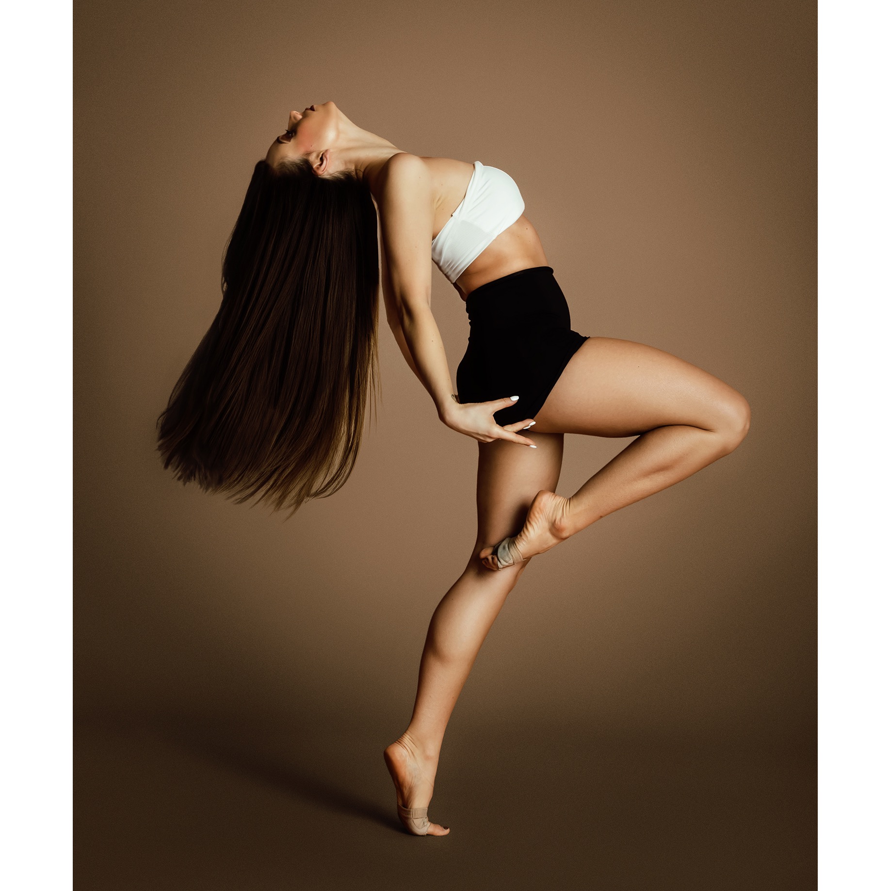 Kobieta w białym topie i czarnych spodenkach w dynamicznej pozie z odchyloną głową i długimi, rozpuszczonymi włosami na jednolitym, brązowym tle.