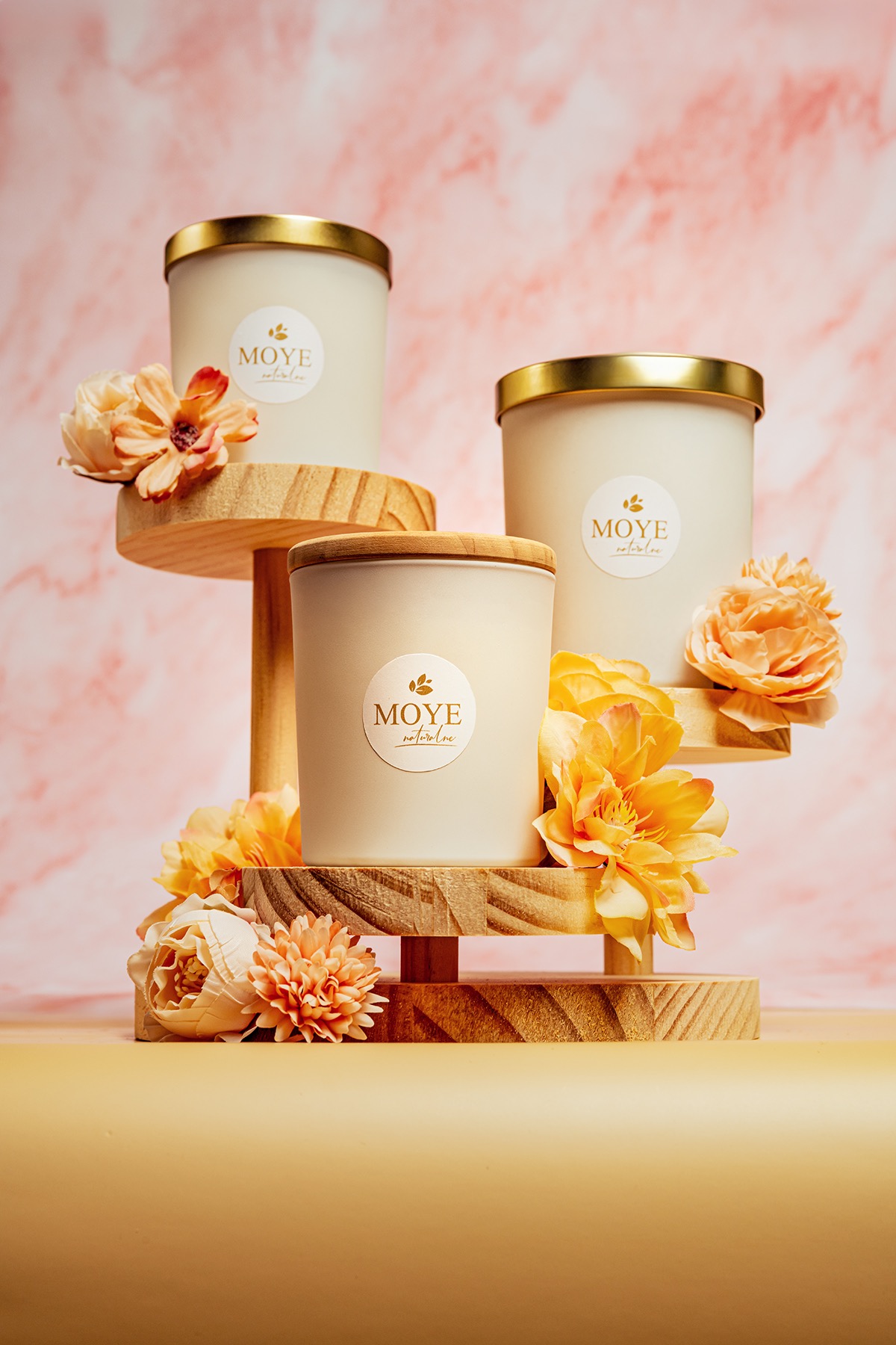 Trzy świece zapachowe marki MOYE natural line na drewnianym stojaku z pomarańczowymi kwiatami na tle marmurkowym.