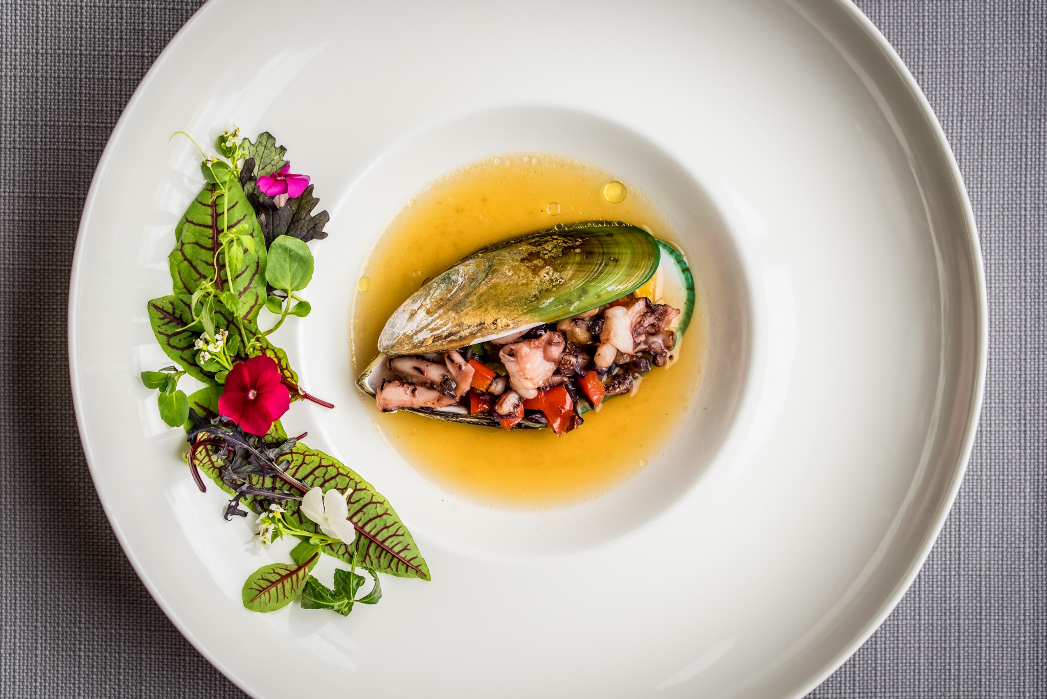 Elegancko podane danie z owoców morza: małża wypełniona ośmiornicą i papryką, pływająca w bulionie, udekorowana świeżymi ziołami i kwiatami na białym talerzu.