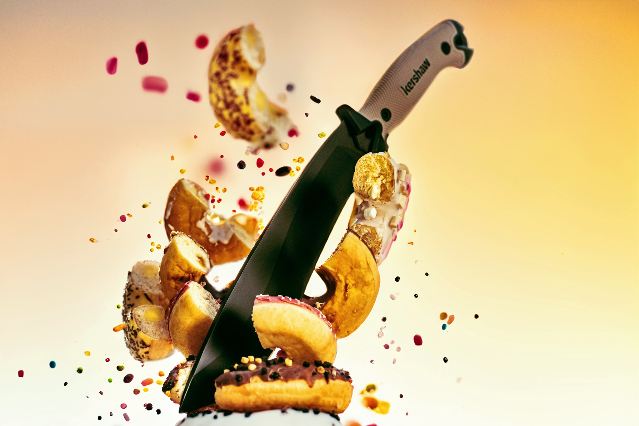 Czarny nóż Kershaw wbity w stos pączków z różnymi polewami i posypkami, unoszące się fragmenty pączków i kolorowe kropki na rozmytym żółto-białym tle.