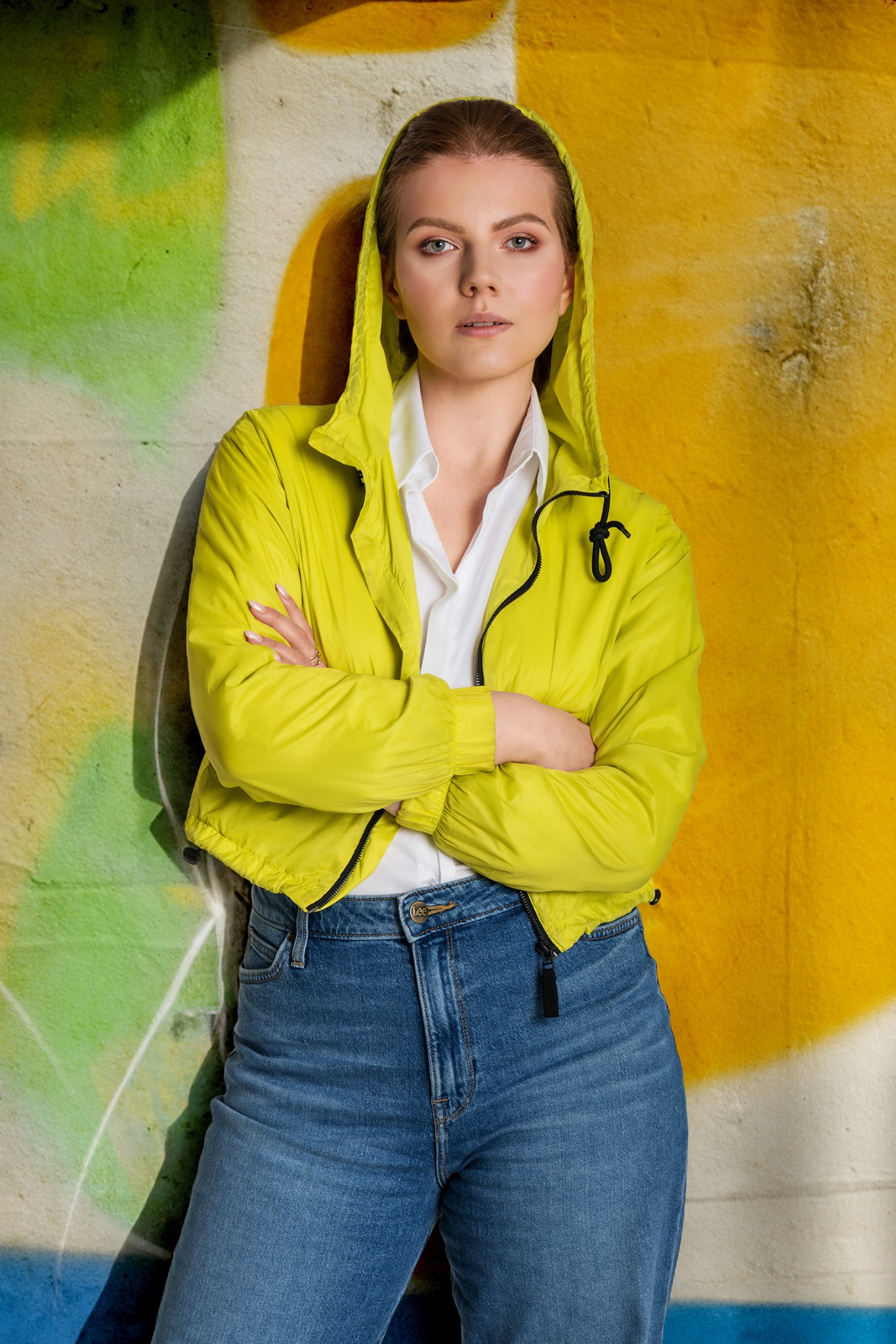 Portret kobiety w limonkowej kurtce z kapturem, opierającej się o ścianę z graffiti w odcieniach zieleni i żółci. Ma skrzyżowane ręce, białą koszulę i dżinsy.