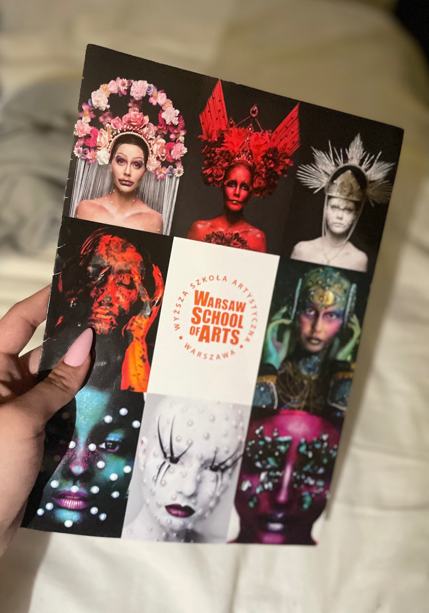 Broszura z okładką prezentującą dziewięć zdjęć artystycznych makijaży i stylizacji twarzy, trzymana w dłoni z różowym paznokciem.