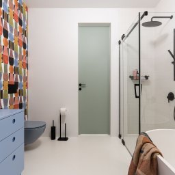 EB PROJEKT EWA BUDNIAK-BEDNARSKA - Nowoczesna łazienka z niebieską szafką pod umywalką, podwieszaną toaletą, kabiną prysznicową ze szklanymi ścianami w czarnych ramach, oraz ścianą z geometryczną tapetą w stylu retro.