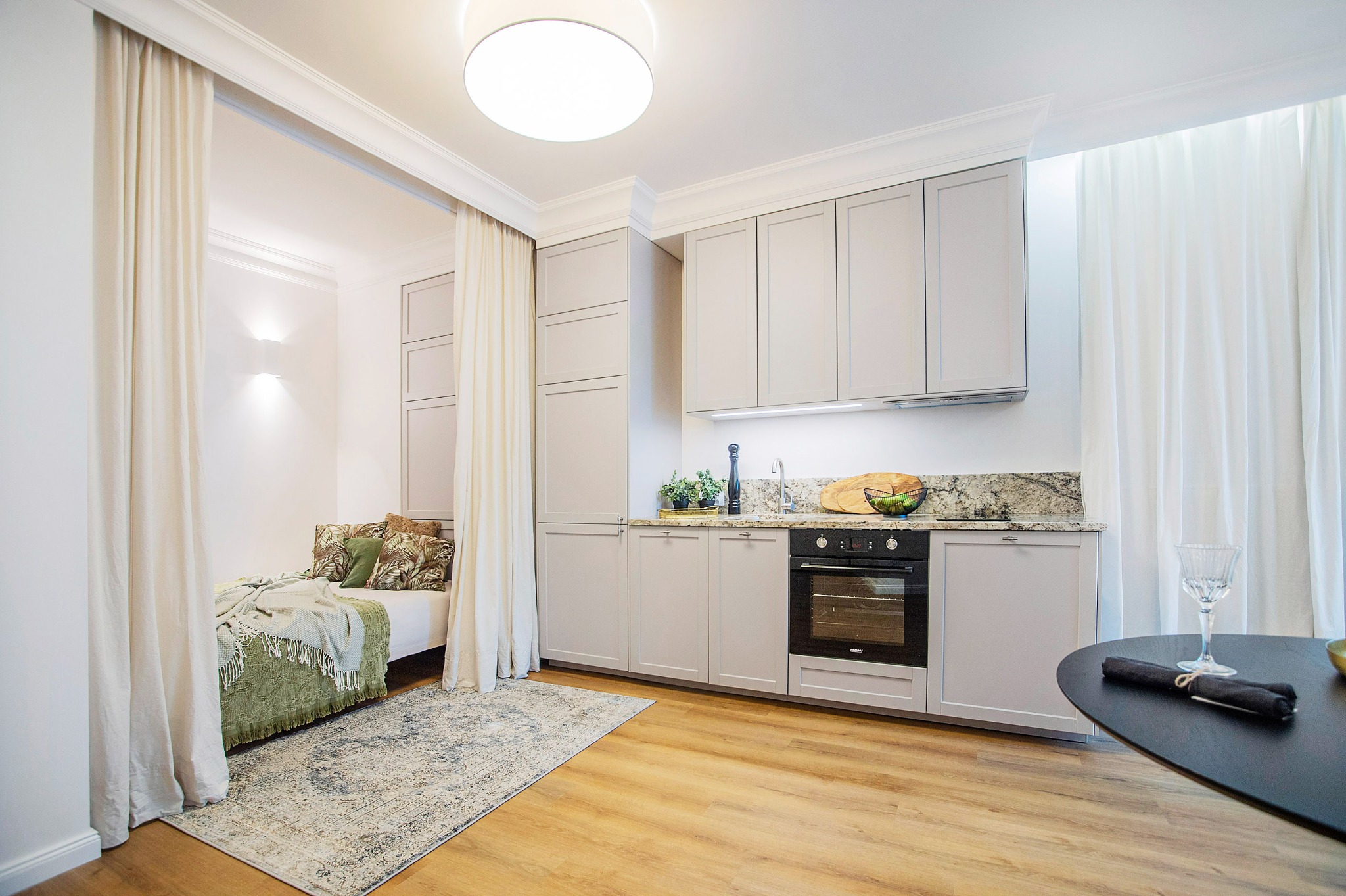 Jasna kawalerka z aneksem kuchennym oddzielonym zasłonami od części sypialnej, wykończona w szarościach z drewnianą podłogą i czarnym okrągłym stołem.