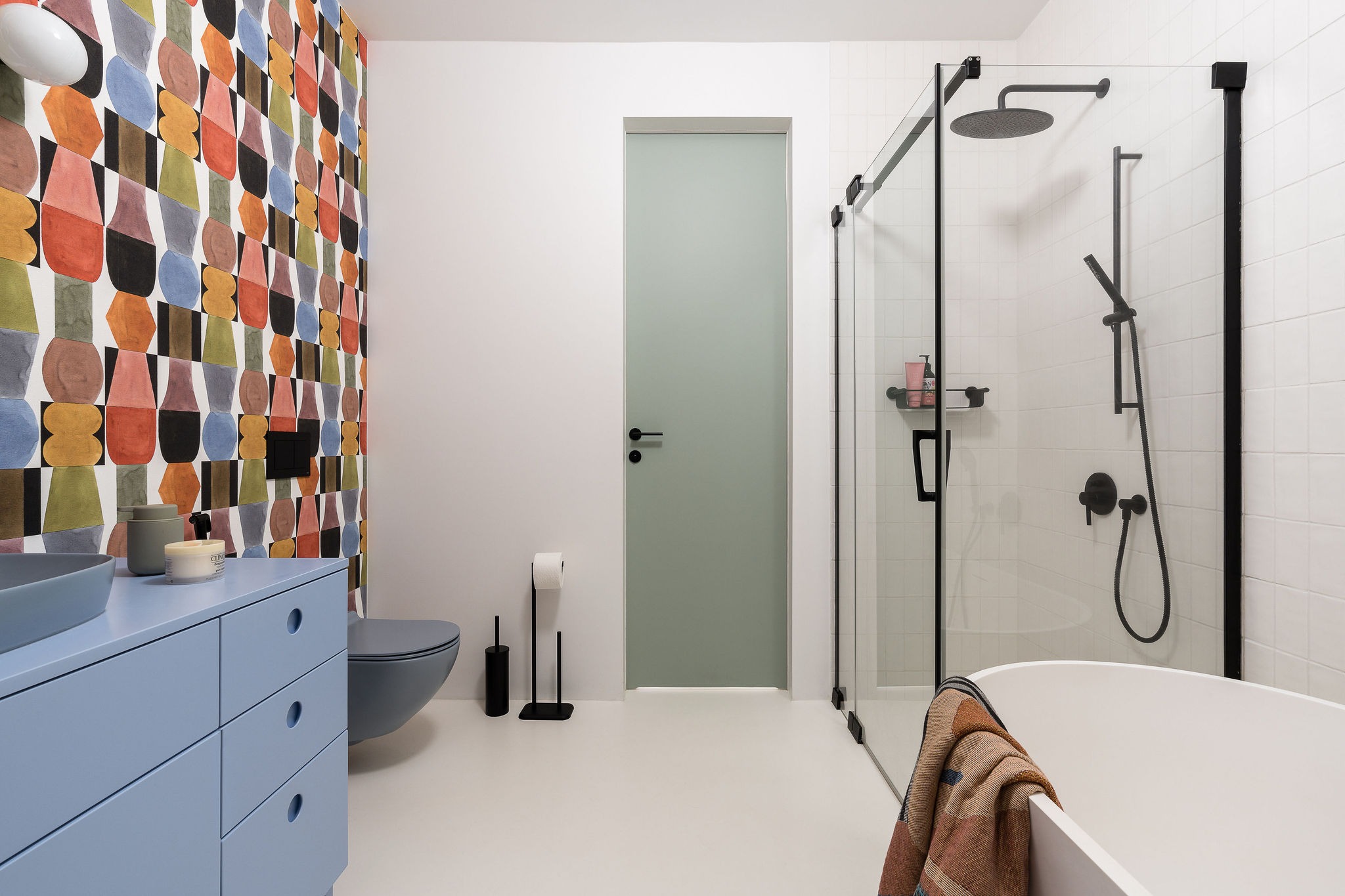 Nowoczesna łazienka z niebieską szafką pod umywalką, podwieszaną toaletą, kabiną prysznicową ze szklanymi ścianami w czarnych ramach, oraz ścianą z geometryczną tapetą w stylu retro.