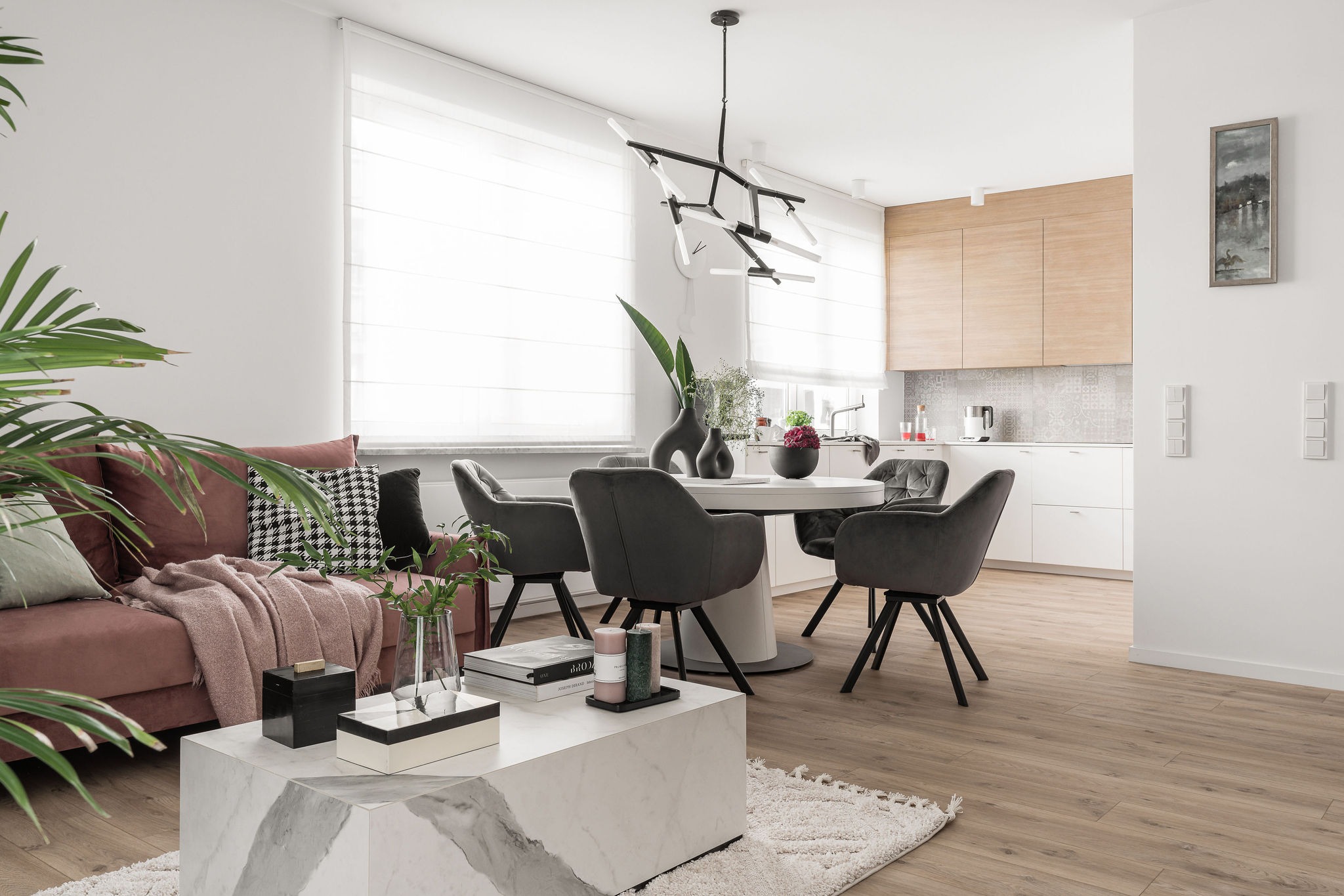 Jasne wnętrze z otwartą kuchnią: pudrowo-różowa sofa z poduszkami w pepitkę, marmurkowy stolik kawowy, okrągły stół z szarymi krzesłami, nowoczesna lampa wisząca i drewniane akcenty w kuchni.