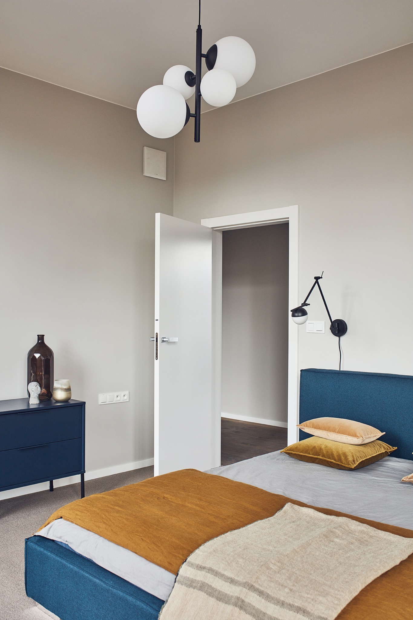 Nowoczesna sypialnia z niebieskim łóżkiem, musztardowymi poduszkami i pledem w paski, uzupełniona czarną lampą wiszącą z białymi kulami i minimalistyczną szafką nocną z dekoracjami.