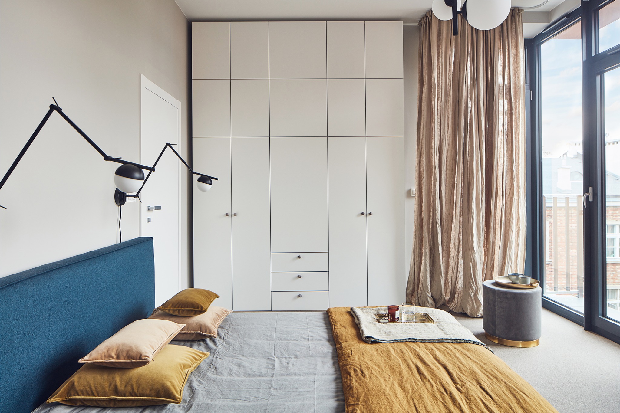 Nowoczesna sypialnia z dużym oknem, zasłonami w kolorze beżowym, łóżkiem z niebieskim wezgłowiem, szarą pościelą i złotymi poduszkami, oraz białą szafą w zabudowie.