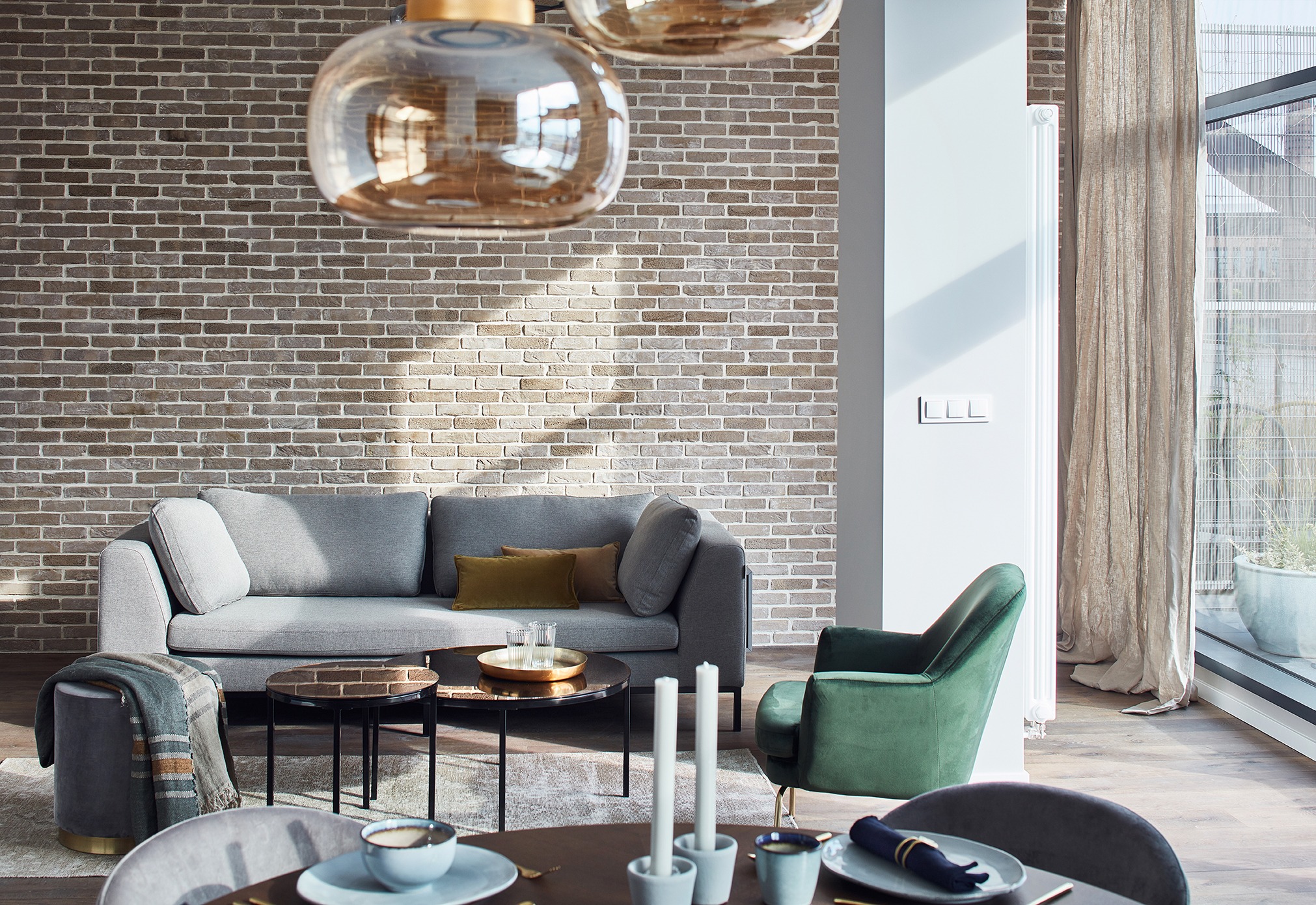 Jasne wnętrze z ceglaną ścianą, szarą sofą z poduszkami, zielonym fotelem i okrągłym stolikiem kawowym z dekoracjami, oświetlone przez wiszące szklane lampy i światło wpadające przez okno.