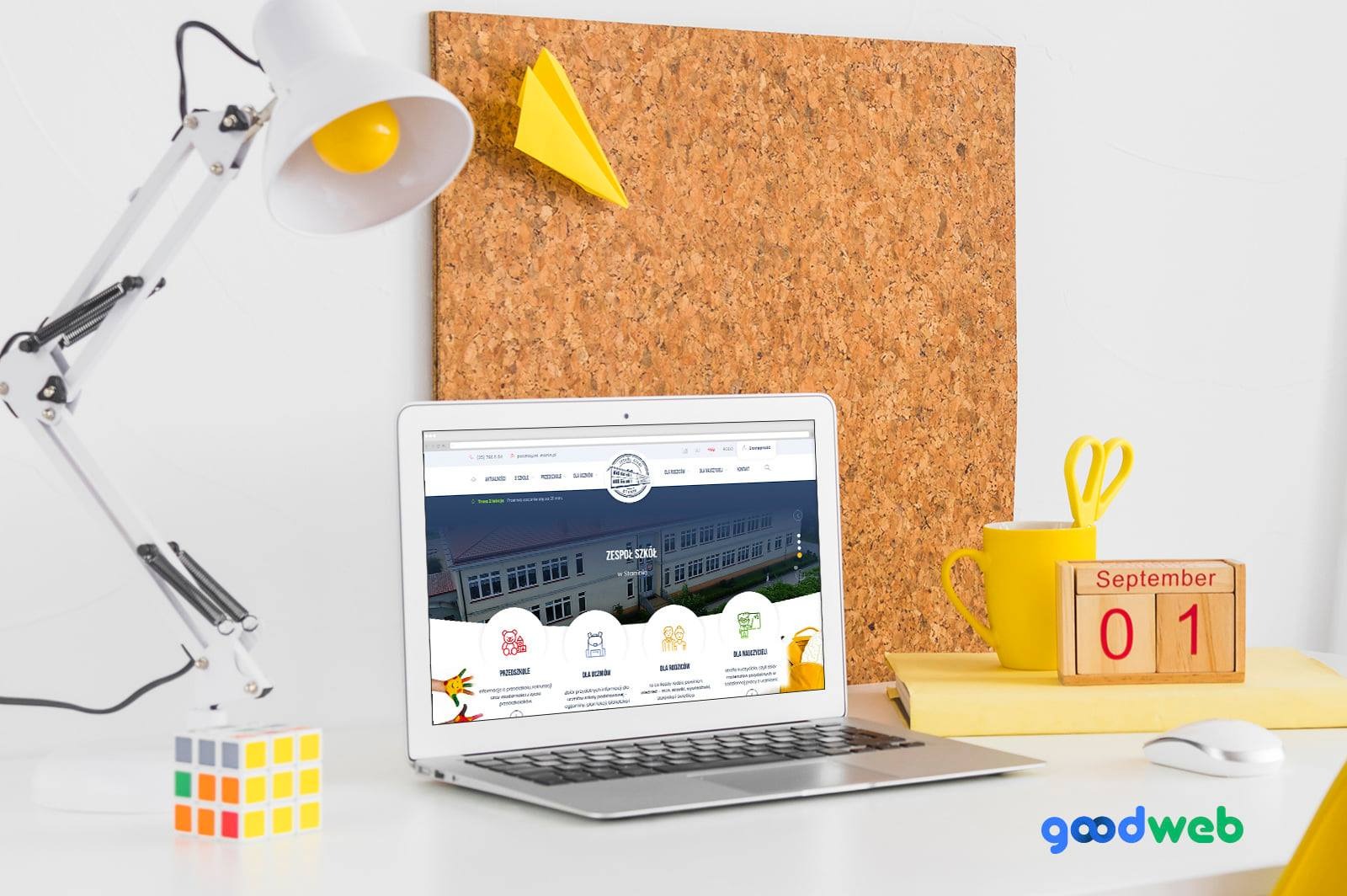 Laptop wyświetlający stronę internetową Zespołu Szkół na biurku z żółtymi akcentami, w tle korkowa tablica z papierowym samolotem.