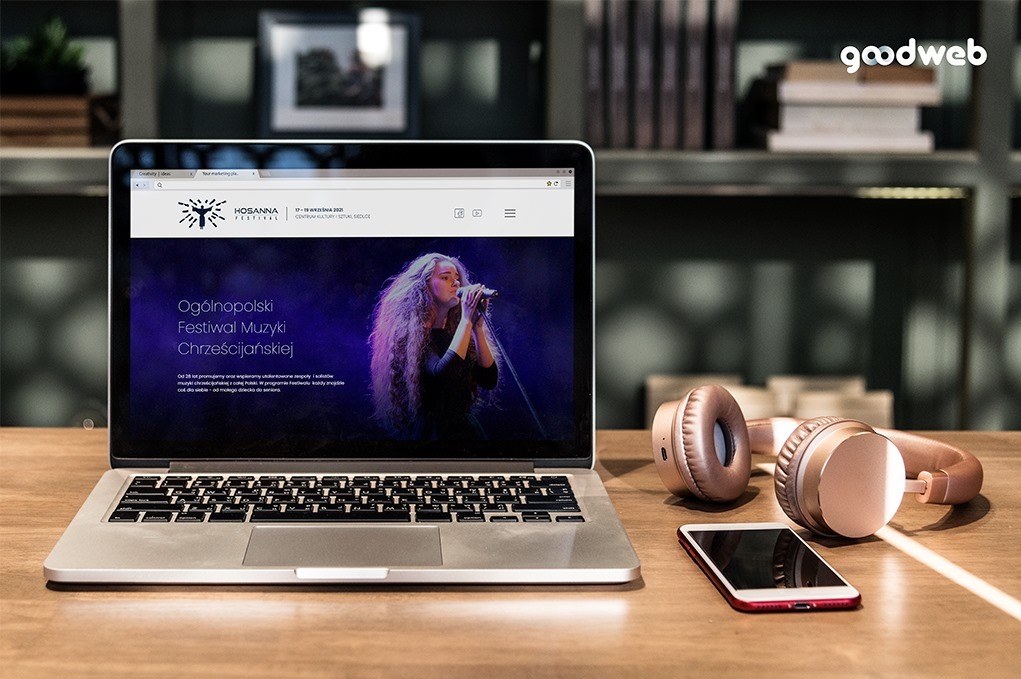 Laptop wyświetlający stronę internetową festiwalu muzycznego, obok słuchawki i smartfon na drewnianym biurku, w tle regał z książkami i dekoracjami.