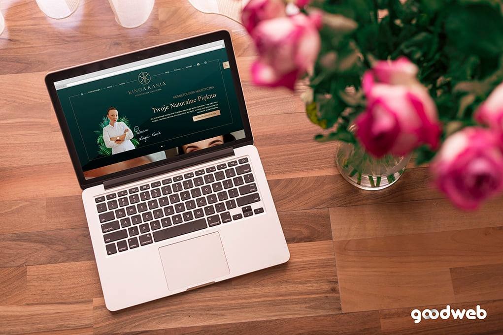 Laptop wyświetla stronę internetową kosmetyczki na drewnianym stole obok wazonu z różowymi różami.