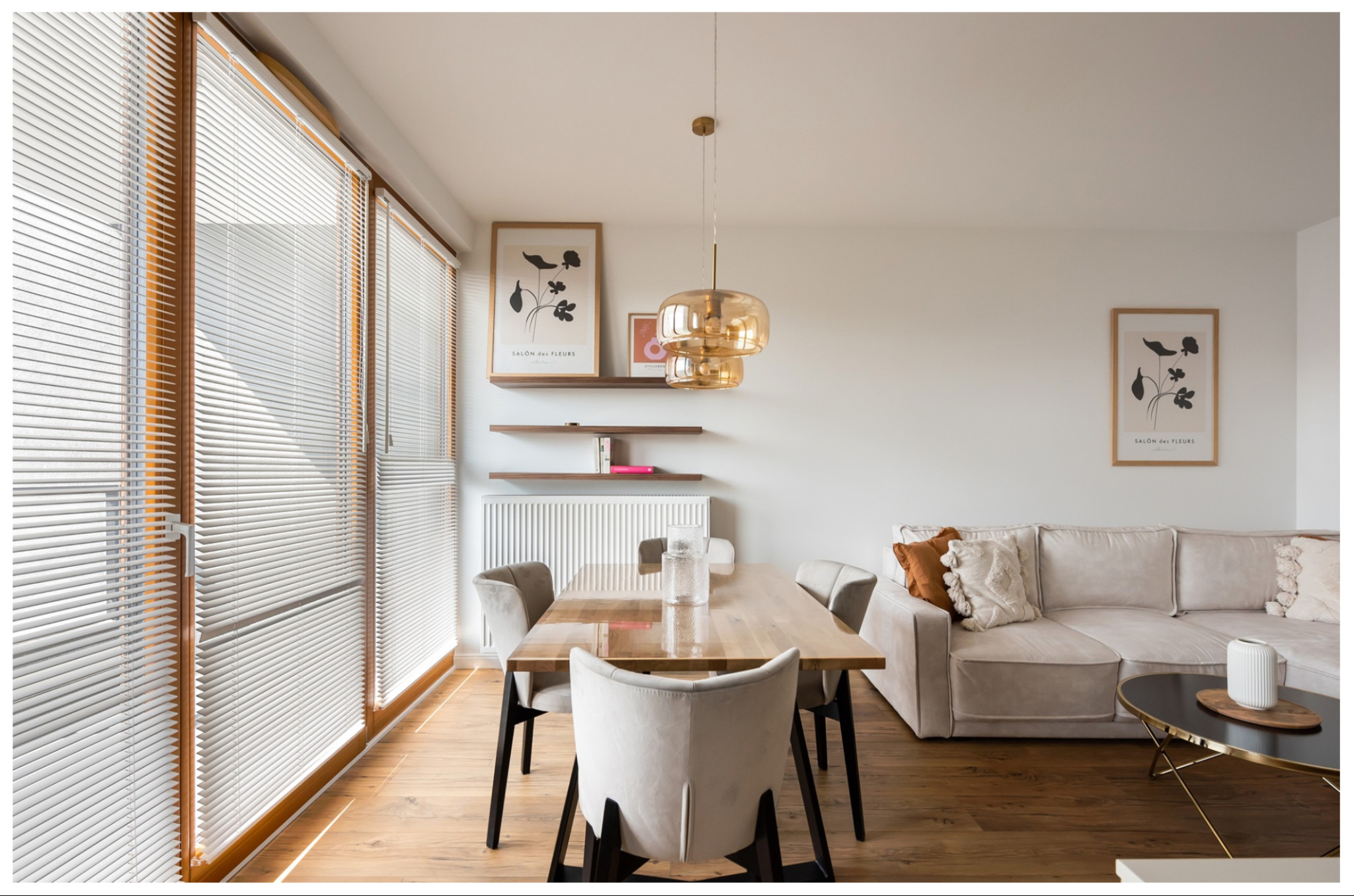Jasne wnętrze mieszkania z drewnianą podłogą, dużym oknem z żaluzjami, stołem z krzesłami, kanapą i złotą lampą wiszącą.