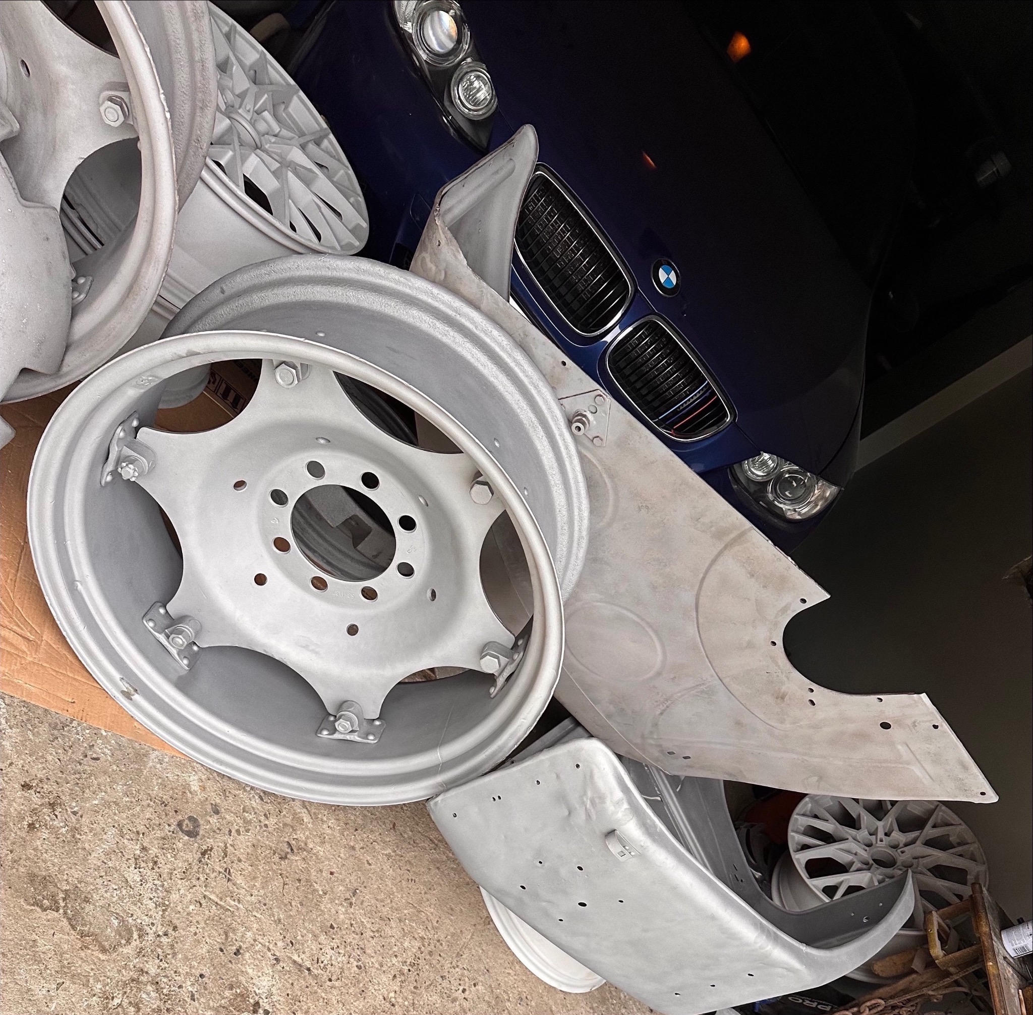 Elementy karoserii i felgi samochodowe po obróbce wstępnej, przygotowane do dalszych prac wykończeniowych, na tle fragmentu ciemnoniebieskiego samochodu BMW.