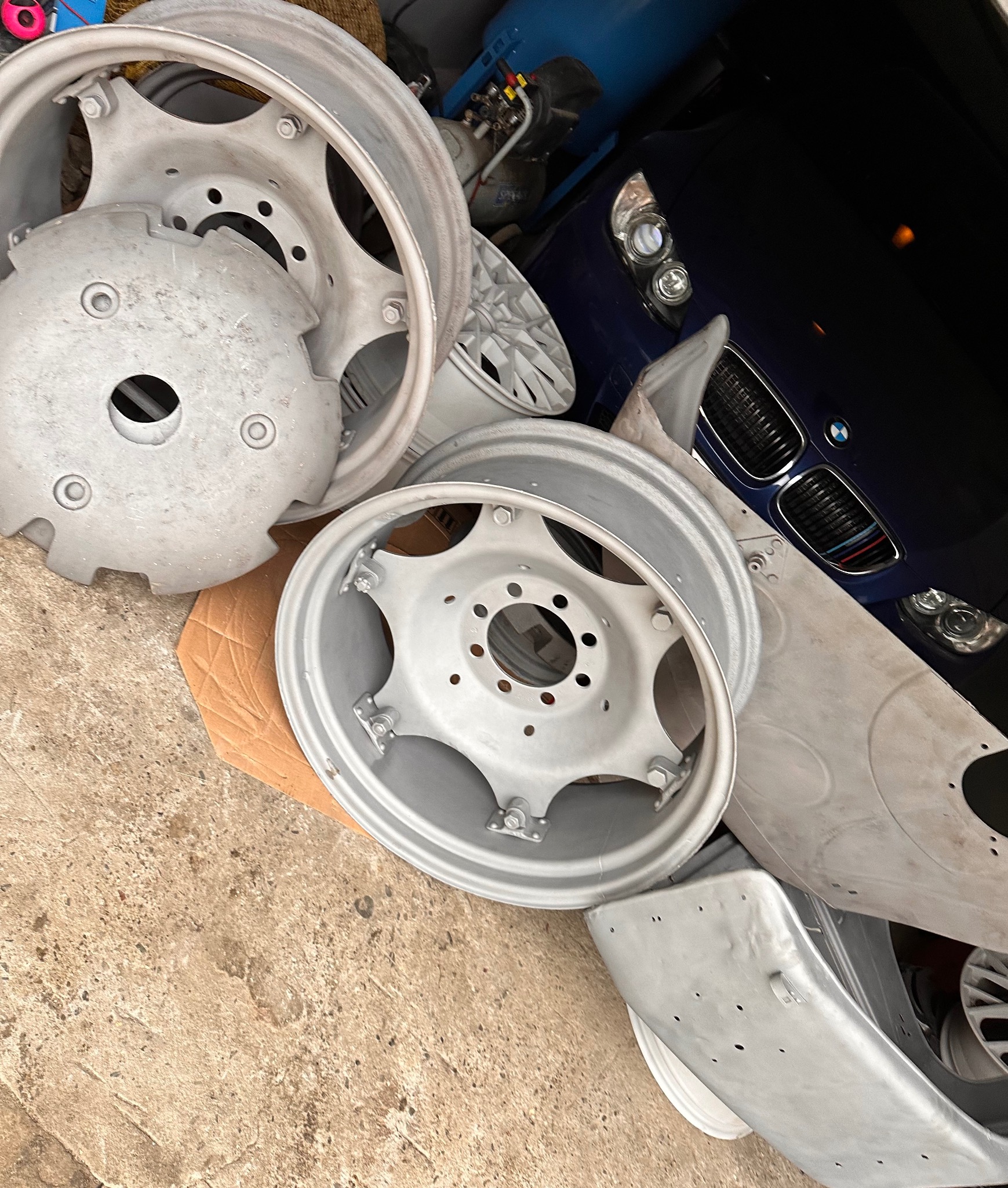 Elementy karoserii i felgi samochodowe, po obróbce przygotowującej do malowania, leżą na betonowej podłodze garażu, w tle widoczny niebieski samochód.