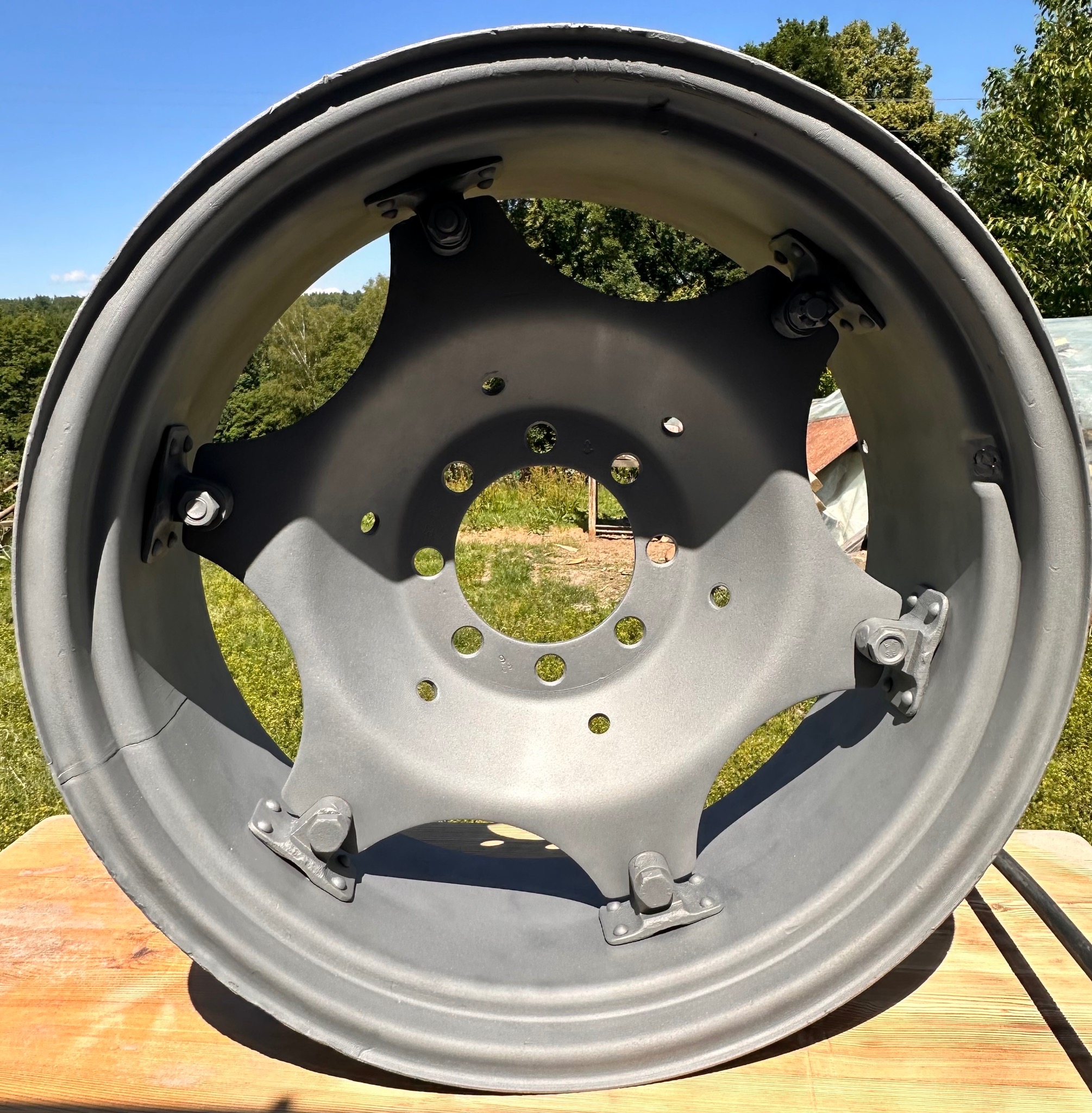Oczyszczona i pomalowana na szaro felga od traktora, leżąca na drewnianym stole z widocznymi śladami użytkowania, na tle zieleni i błękitnego nieba.