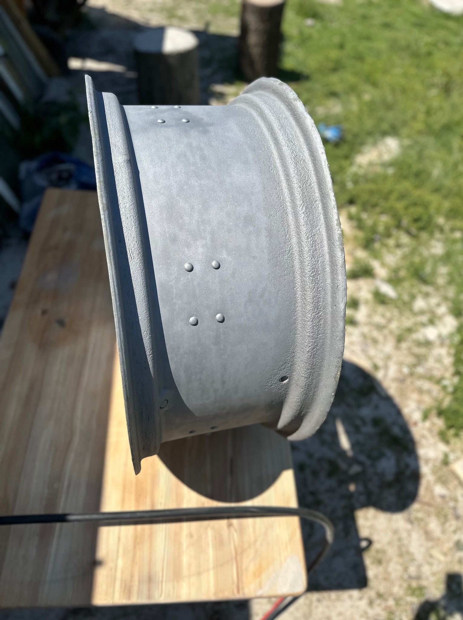 Szara felga samochodowa po obróbce, leżąca na drewnianym stole w plenerze, widoczne ślady po procesie czyszczenia i przygotowania do malowania.