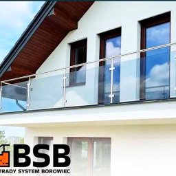 BSB Mateusz Borowiec - Balkon z nowoczesną szklaną balustradą ze słupkami ze stali nierdzewnej, widok z dołu na tle błękitnego nieba i białej elewacji domu z brązowymi oknami, w rogu widoczne logo firmy.