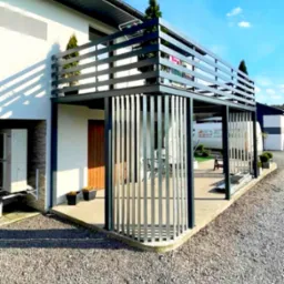 Nowoczesna, dwupoziomowa konstrukcja z metalu w kolorze antracytowym, pełniąca funkcję tarasu i osłony, z pionowymi elementami dekoracyjnymi, przylegająca do białego budynku z elementami kamiennymi...