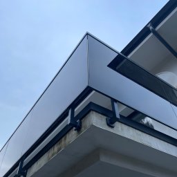 BSB Mateusz Borowiec - Szklana balustrada balkonowa z czarnymi metalowymi elementami mocującymi, widok z dołu na tle jasnego nieba.