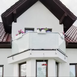 BSB Mateusz Borowiec - Balkon z nowoczesną, szklaną konstrukcją w Kielcach, ozdobiony wiszącymi i stojącymi donicami z kwiatami, na tle białej elewacji budynku z brązowym dachem.