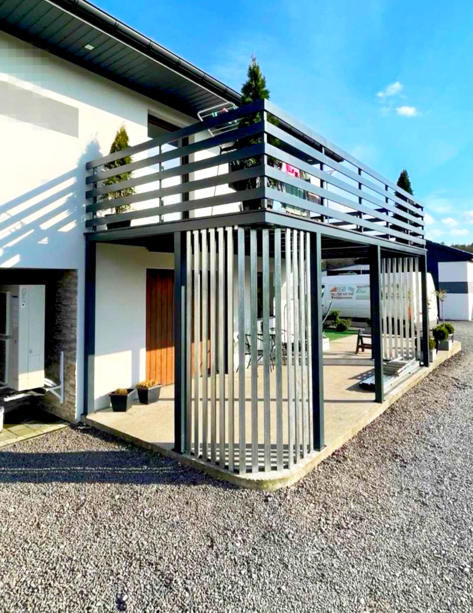 Nowoczesna, dwupoziomowa konstrukcja z metalu w kolorze antracytowym, pełniąca funkcję tarasu i osłony, z pionowymi elementami dekoracyjnymi, przylegająca do białego budynku z elementami kamiennymi...