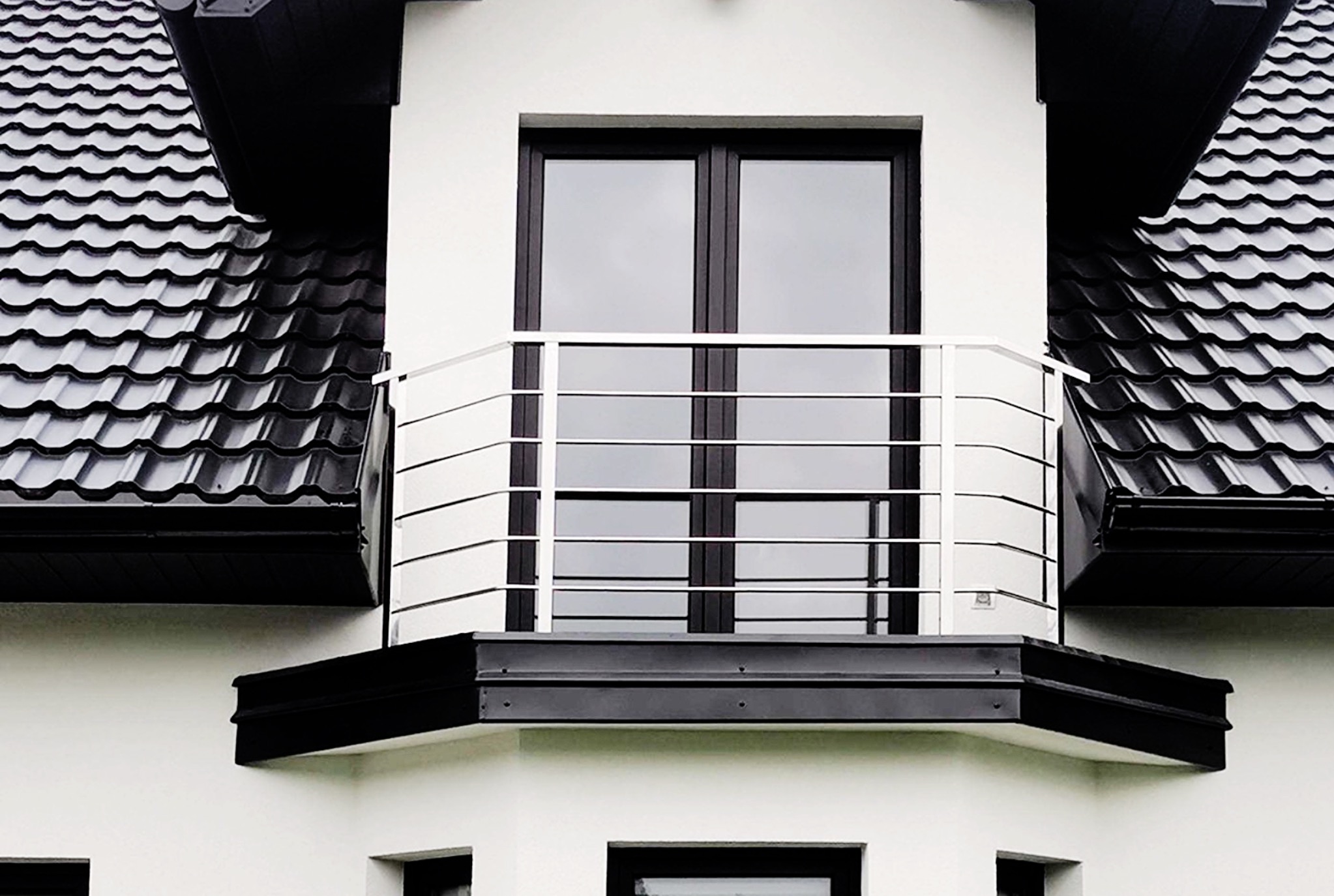 Nowoczesny balkon z białą elewacją i czarną stolarką okienną, zabezpieczony prostą, poziomą konstrukcją z cienkich, metalowych prętów, na tle czarnego, błyszczącego dachu z dachówki ceramicznej.