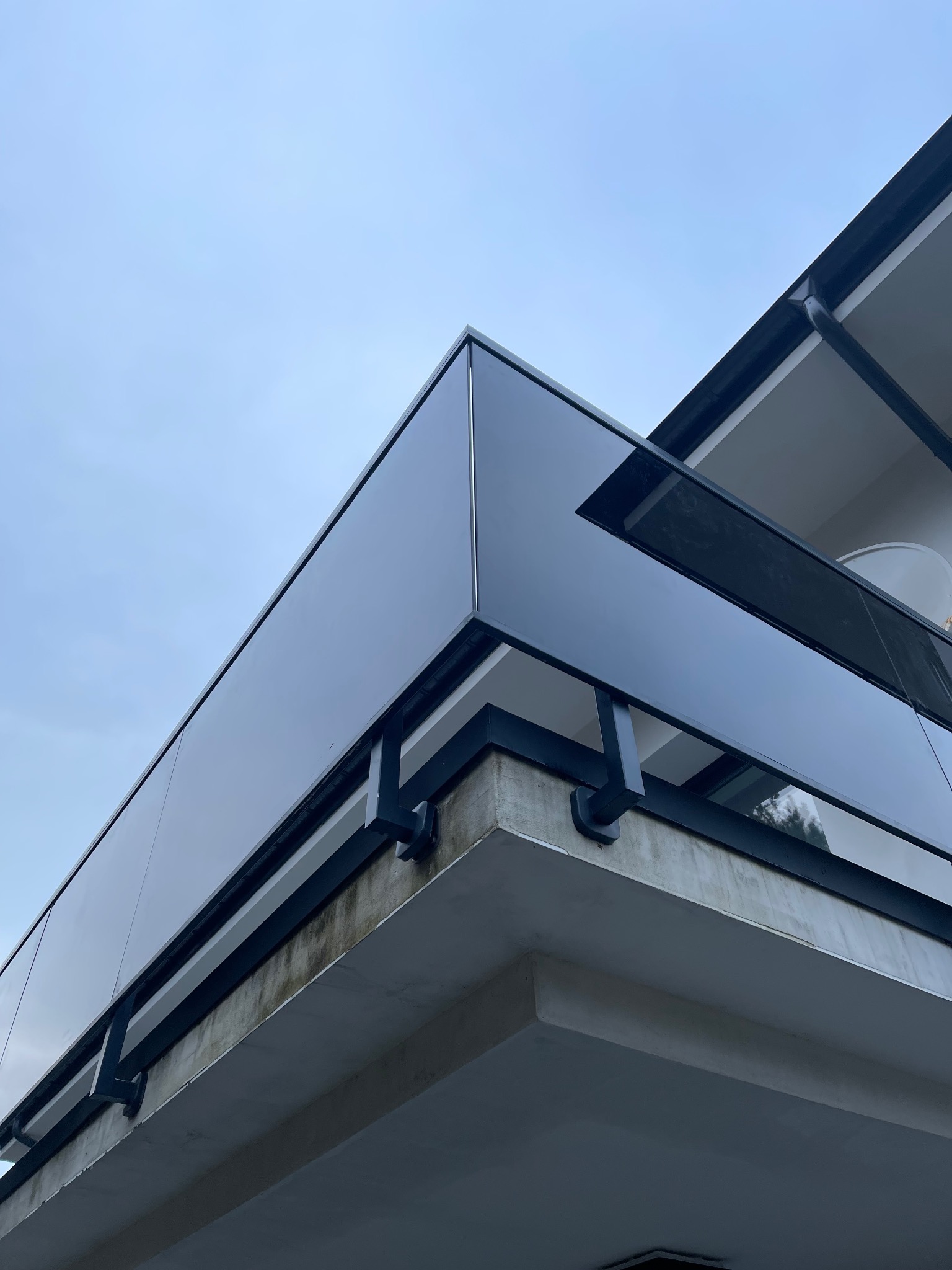 Szklana balustrada balkonowa z czarnymi metalowymi elementami mocującymi, widok z dołu na tle jasnego nieba.