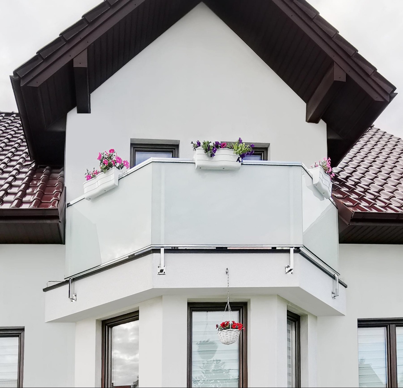 Balkon z nowoczesną, szklaną konstrukcją w Kielcach, ozdobiony wiszącymi i stojącymi donicami z kwiatami, na tle białej elewacji budynku z brązowym dachem.