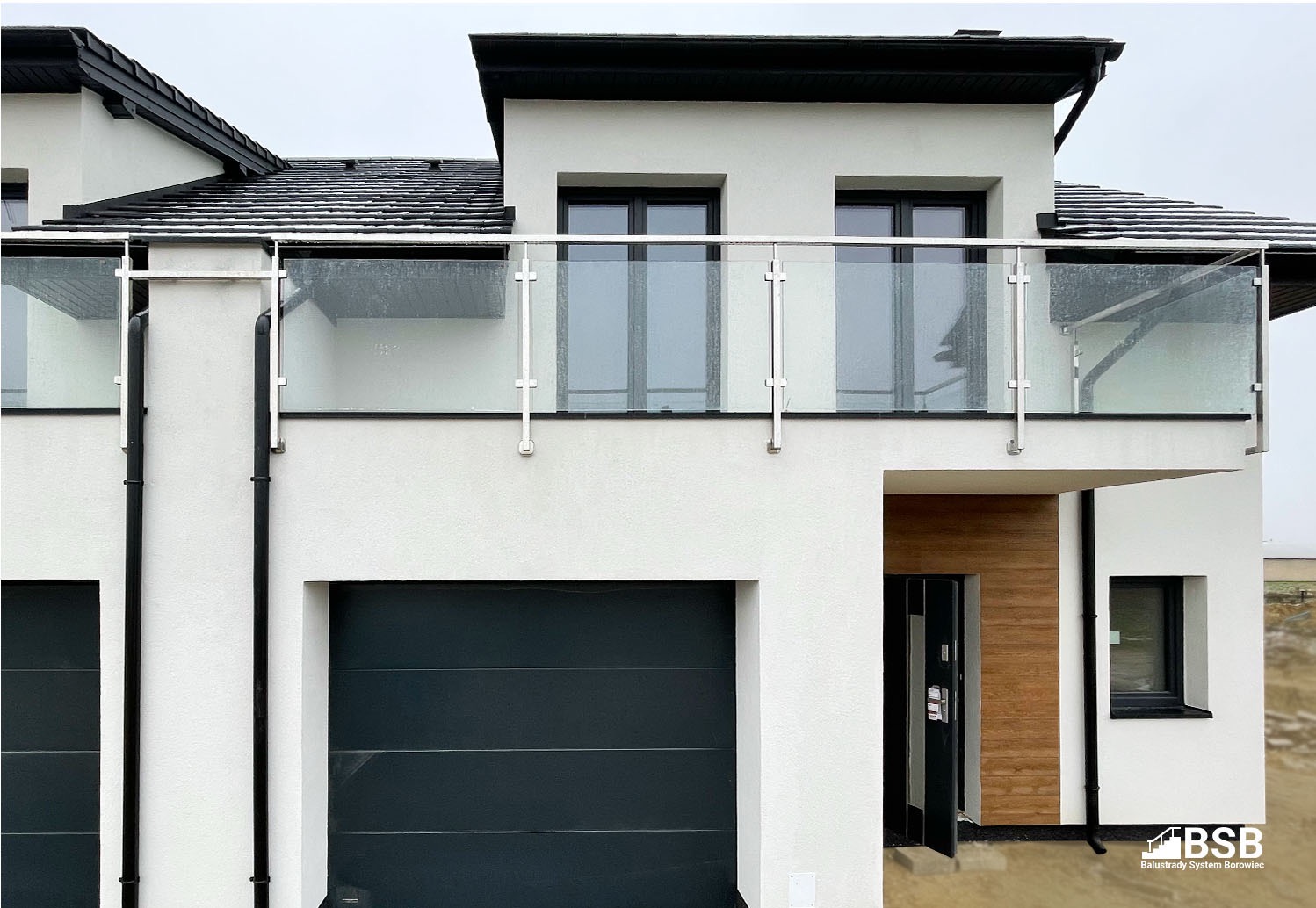 Nowoczesny dom z białymi ścianami i czarnymi detalami, balkon z oszkloną balustradą na pierwszym piętrze, garaż z ciemną bramą, wejście z drewnianym akcentem, widoczny znak firmowy w prawym dolnym...