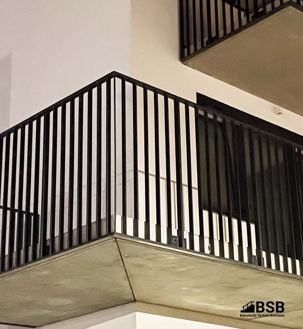 Czarne, pionowe pręty zabezpieczające balkon na tle białej ściany, widok z dołu, logo firmy w prawym dolnym rogu.