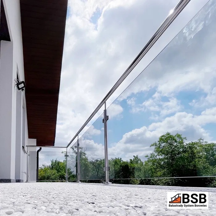 Balkon z nowoczesną, przezroczystą balustradą szklaną z elementami ze stali nierdzewnej, podłoga wyłożona białym kamieniem, widok na zieleń i błękitne niebo, w rogu widoczne logo firmy.