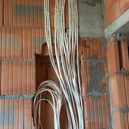 K&A INSTAL BUD - Białe kable elektryczne ułożone w peszlach, prowadzone po ceglanej ścianie w surowym stanie, część kabli zwisająca luźno ze ściany, zabezpieczona żółtą taśmą.