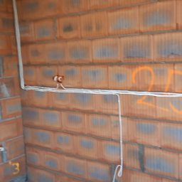 K&A INSTAL BUD - Surowe ściany z czerwonej cegły z widocznymi kablami elektrycznymi w białej izolacji, wychodzącymi z puszek instalacyjnych. Na ścianie pomarańczowe oznaczenia.