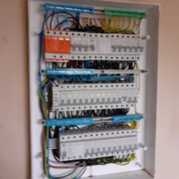 Rozdzielnica elektryczna w białej obudowie, widoczne zabezpieczenia różnicowoprądowe i nadprądowe, przewody ułożone wewnątrz skrzynki, zamontowana na ścianie w trakcie prac wykończeniowych.