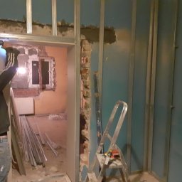 K&A INSTAL BUD - Remont pokoju: montaż metalowej konstrukcji ścianki działowej, widoczne profile, drabina, częściowo usunięta ściana z cegły i pracownik z wiertarką.