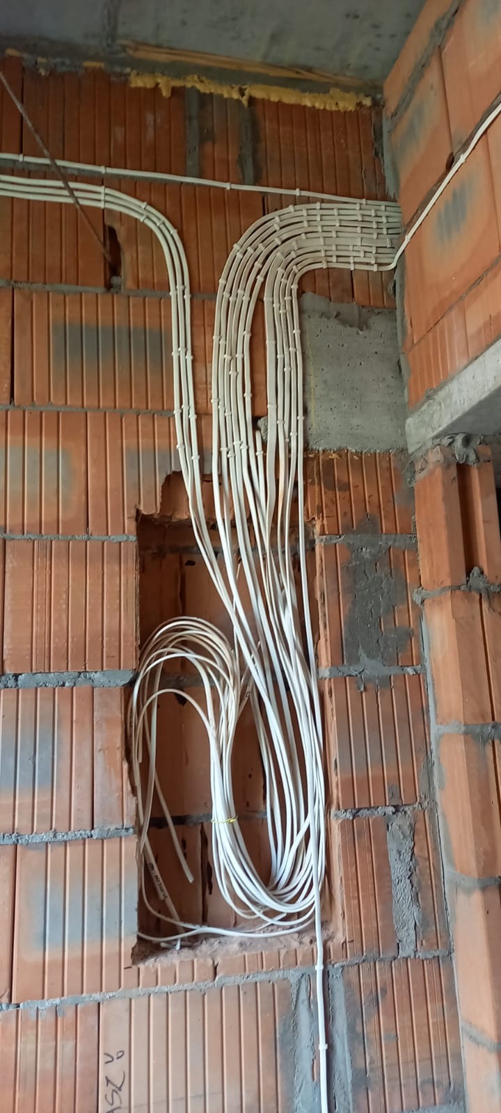 Białe kable elektryczne ułożone w peszlach, prowadzone po ceglanej ścianie w surowym stanie, część kabli zwisająca luźno ze ściany, zabezpieczona żółtą taśmą.