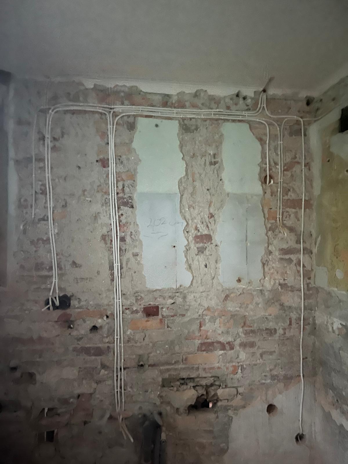 Białe kable elektryczne prowadzone po surowej, ceglanej ścianie w trakcie remontu, z widocznymi fragmentami starego tynku i śladami po instalacjach.