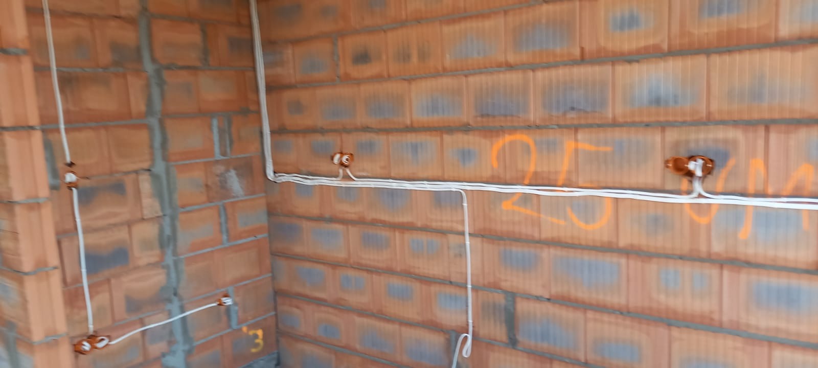 Surowe ściany z czerwonej cegły z widocznymi kablami elektrycznymi w białej izolacji, wychodzącymi z puszek instalacyjnych. Na ścianie pomarańczowe oznaczenia.