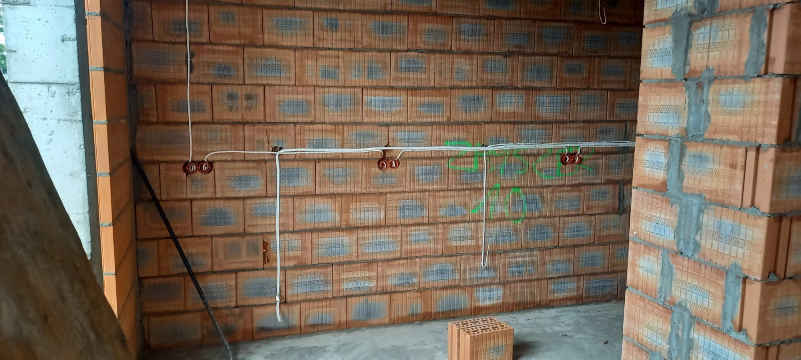 Wewnętrzna ściana budynku w trakcie budowy z widocznymi przewodami elektrycznymi i puszkami w kolorze ceglastym, zielone oznaczenia na ścianie.