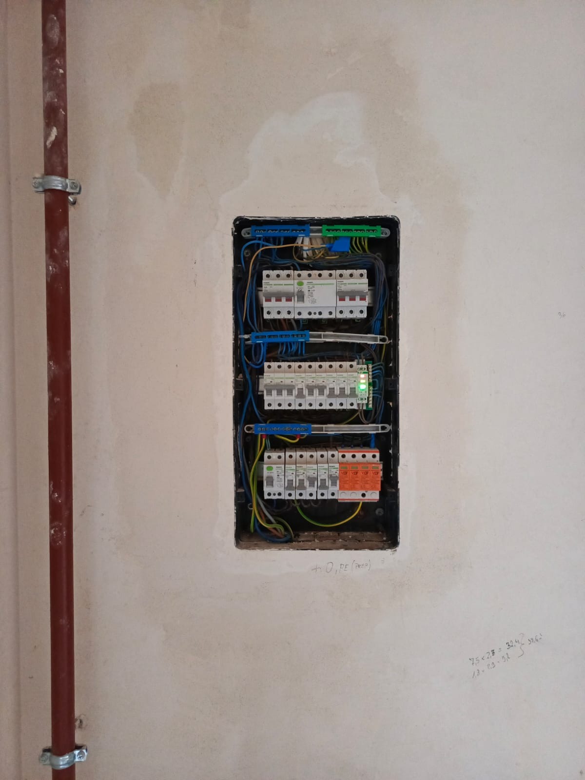 Rozdzielnica elektryczna z bezpiecznikami, przewodami i listwami zaciskowymi zamontowana w ścianie o surowym wykończeniu, widoczny fragment instalacji rurowej po lewej stronie.