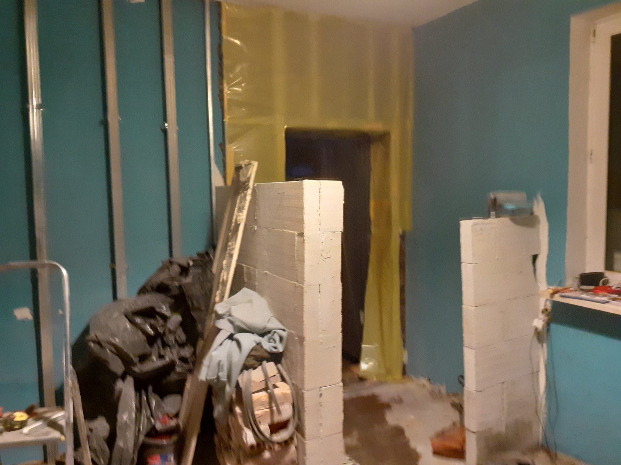Wnętrze w trakcie remontu: ściany w kolorze turkusowym, metalowe profile, fragment ściany z białych bloczków, podłoga zabezpieczona folią, widoczne worki z odpadami i drabina.