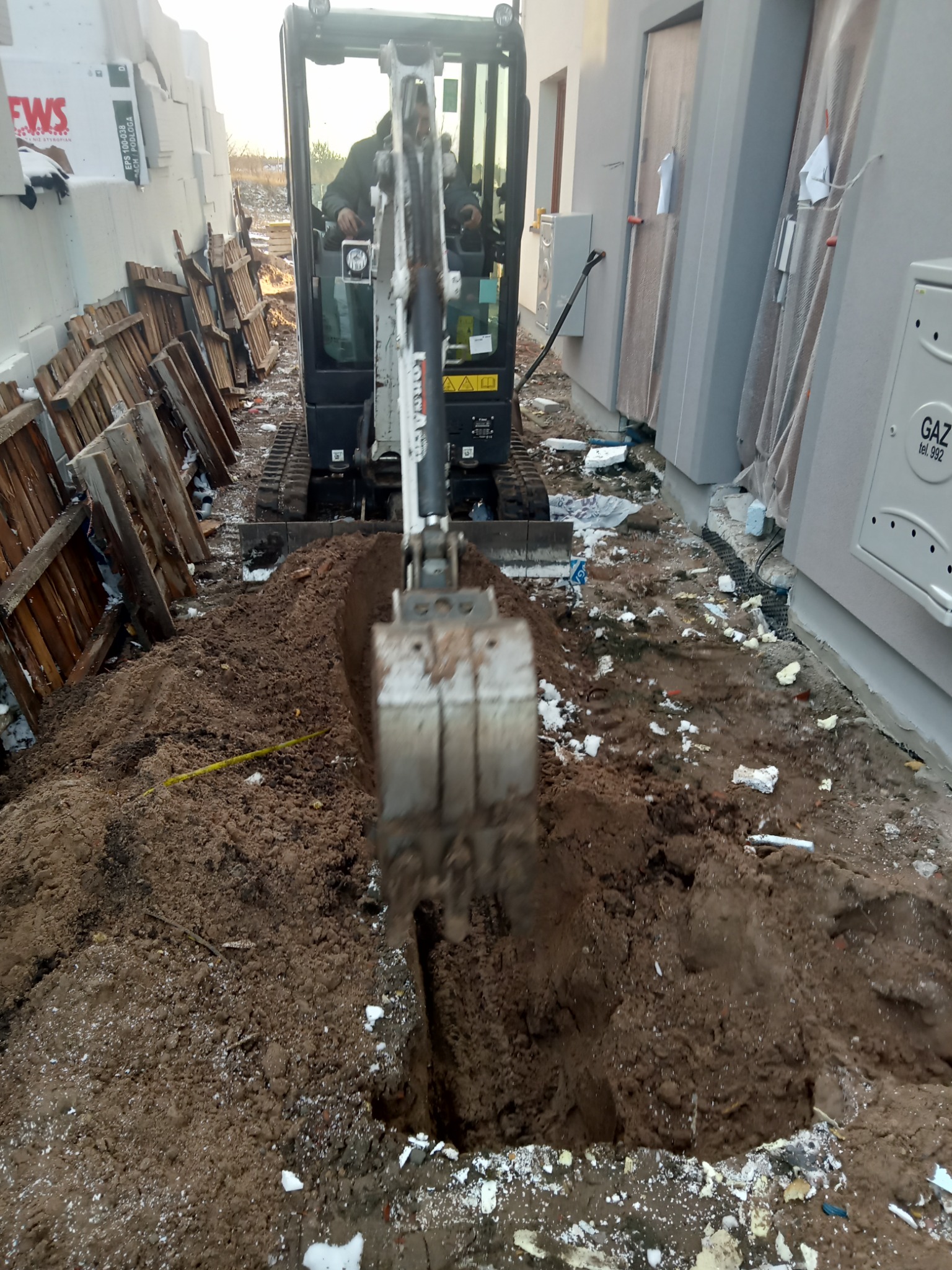 Mini koparka Bobcat wykonuje wykop w ziemi wzdłuż ściany budynku, widoczne palety i styropian.