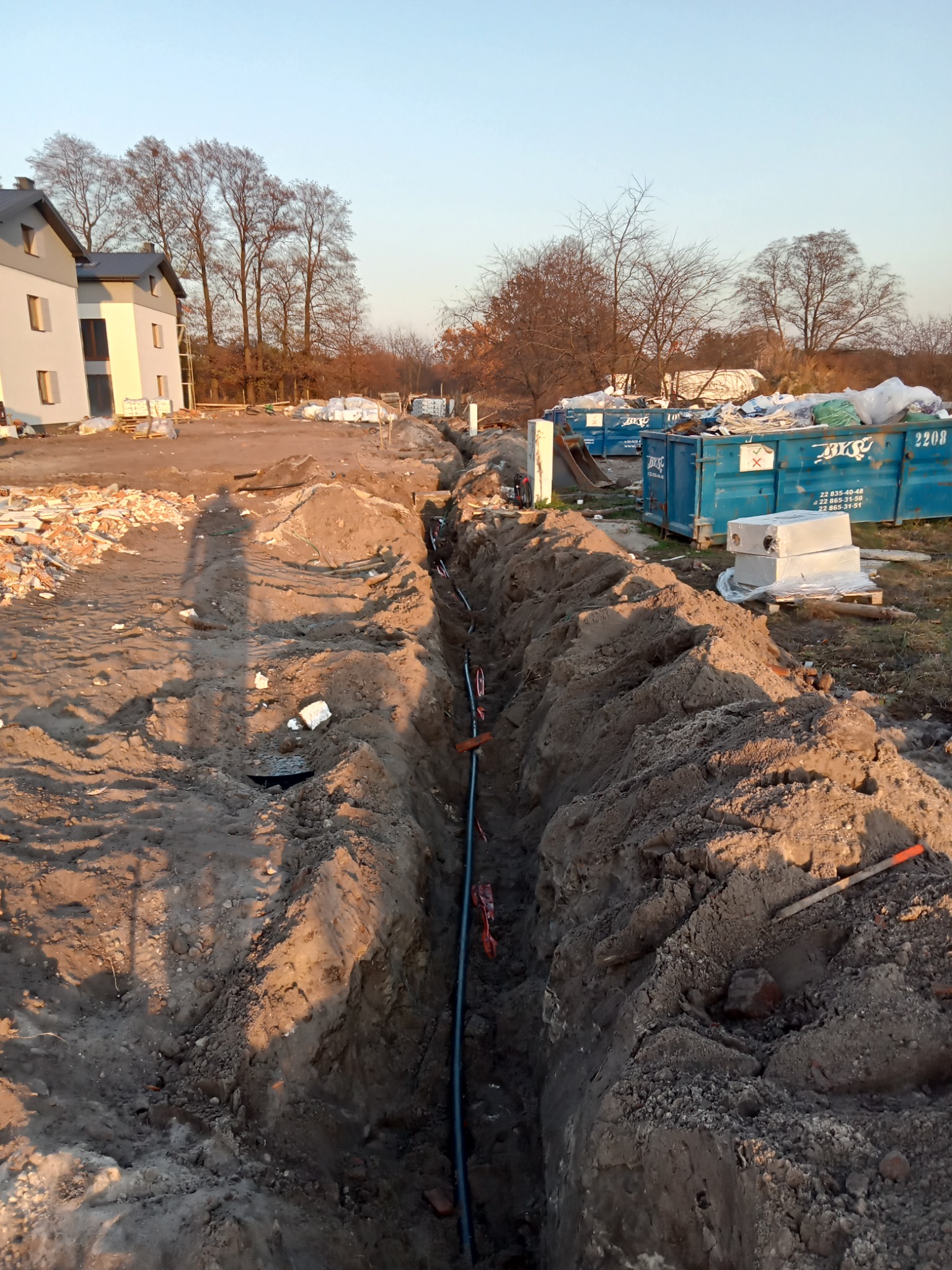 Wykop w ziemi z ułożonym przewodem elektrycznym, widoczne zabezpieczenia kabli, w tle nowo budowane domy i kontenery na gruz.