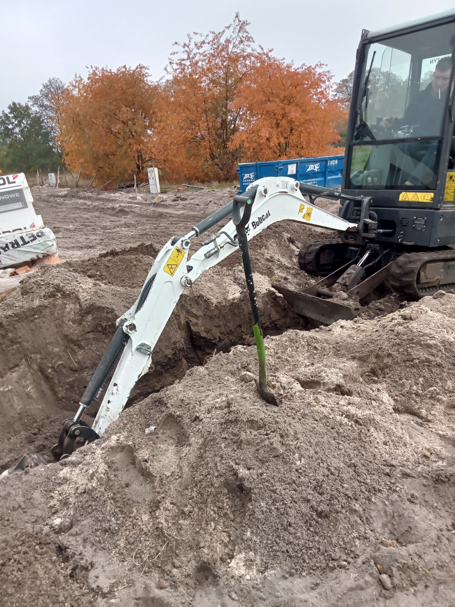 Mini koparka Bobcat w trakcie prac ziemnych, wykop pod instalacje na placu budowy, widoczna łopata z zielonym uchwytem i operator w kabinie.