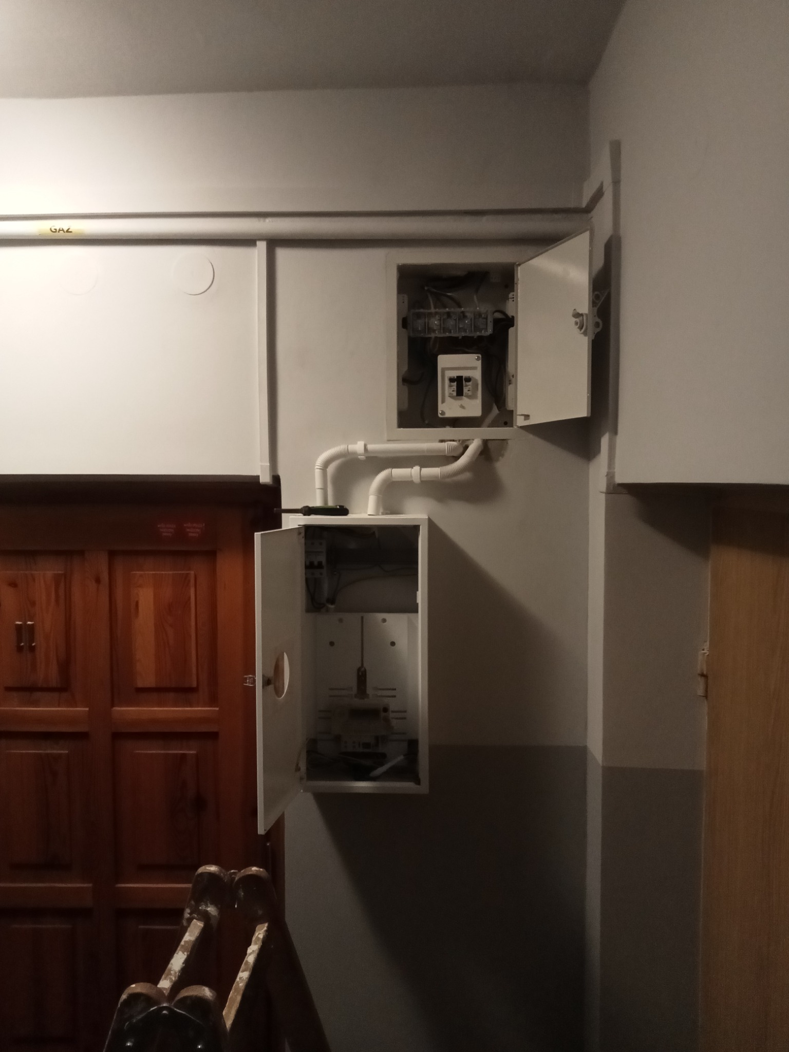 Dwa otwarte panele elektryczne na ścianie, z widocznymi przewodami i elementami instalacji, rury gazowe u góry, drewniane drzwi po bokach, fragment drabiny w dolnej części kadru.