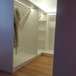 Omeblowani Mariusz Jastrzębski - Wnętrze narożnej, białej garderoby z oświetleniem LED, widoczne ubrania na wieszakach i puste półki, parkiet na podłodze.